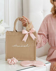 Woman in pink robe holding a beige tote bag with 'Hannah' on it, next to a pink scrunchie and eye mask.