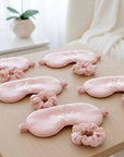 Set of pink eye masks with matching scrunchies on a wooden surface, with a blurred background.