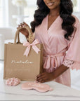 Woman in a pink robe holding a personalized gift bag with matching satin robe, scrunchie and sleep mask set against a blurred indoor background.