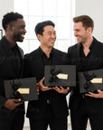 Three groomsmen in suits holding all black gift boxes with gold accents personalized by bossy creations in a bright room.
