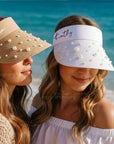 Two women, a bride to be and a bridesmaid wearing personalized visors on a beach with ocean in the background