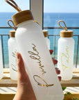 glass water bottles with personalized gold text and wooden caps held by a hand, with a scenic background for bridesmiad gifts.