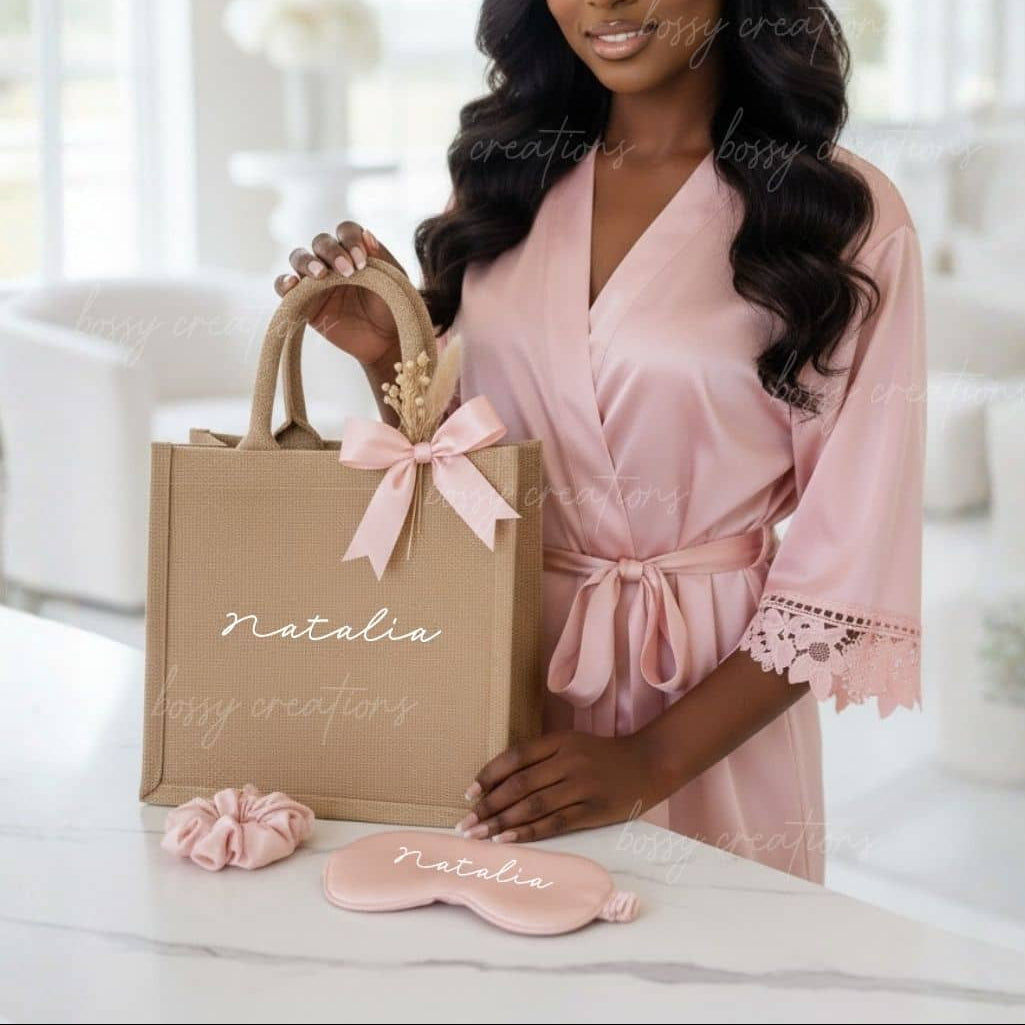 Woman in a pink robe holding a personalized gift bag with matching satin robe, scrunchie and sleep mask set against a blurred indoor background.