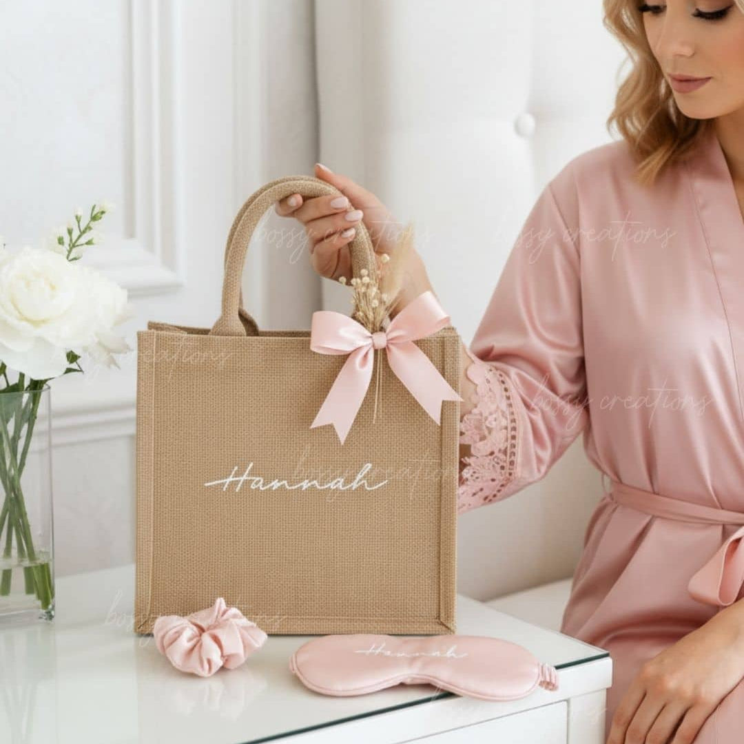 Woman in pink robe holding a beige tote bag with 'Hannah' on it, next to a pink scrunchie and eye mask.