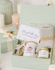 Sage green and pink christmas self care winter Gift box with candle, personalized gingerbread mug and socks on a decorative table with flowers and candles.