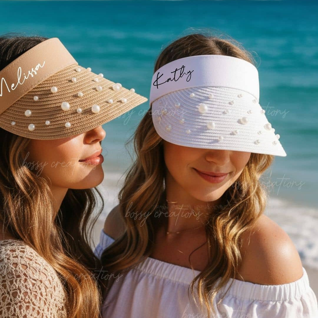 Two women, a bride to be and a bridesmaid wearing personalized visors on a beach with ocean in the background