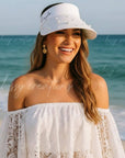 Woman in a white dress and personalized visor standing on a beach with ocean in the background