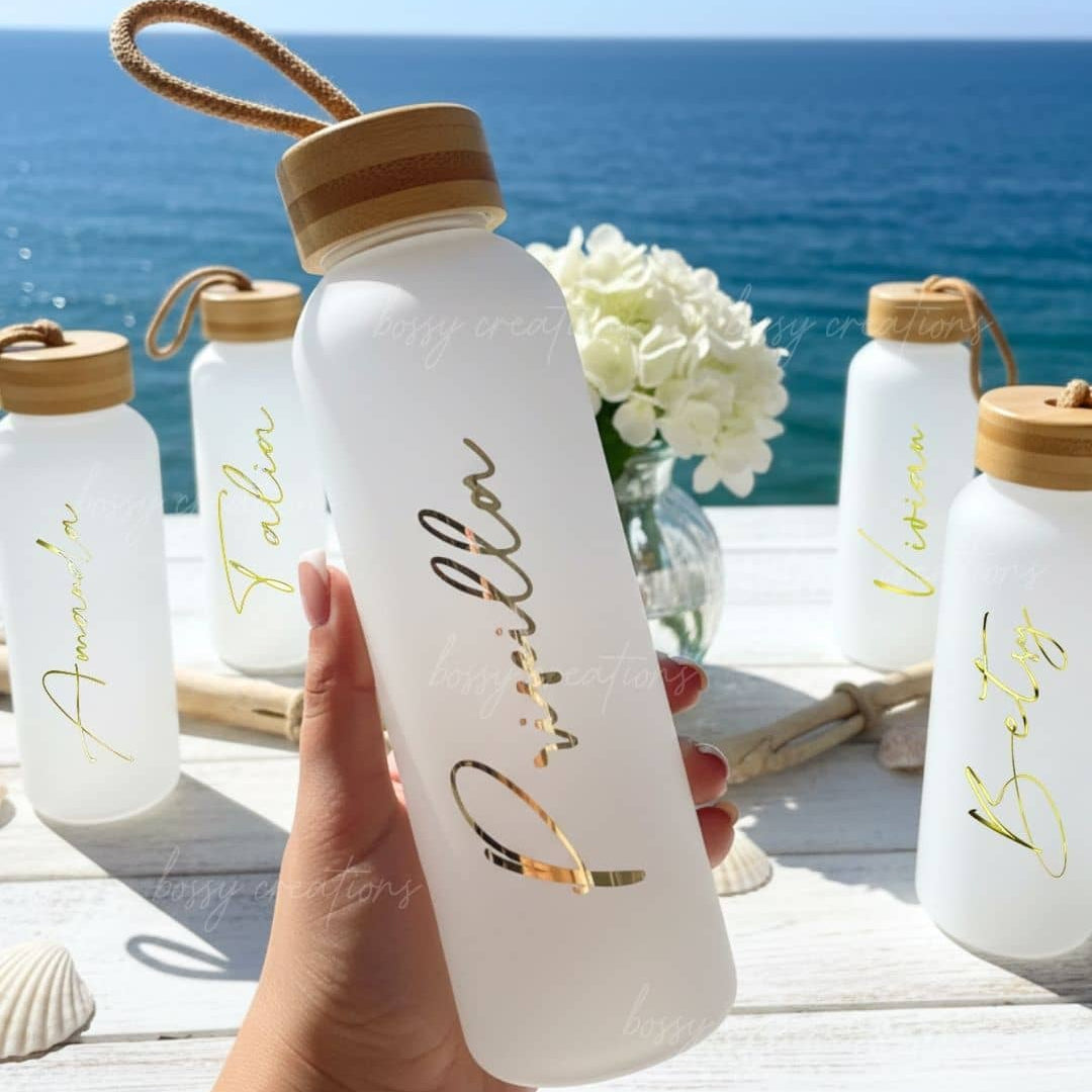White glass water bottle with personalized gold text held by a hand, set against a scenic ocean backdrop during a beach bachelorette.