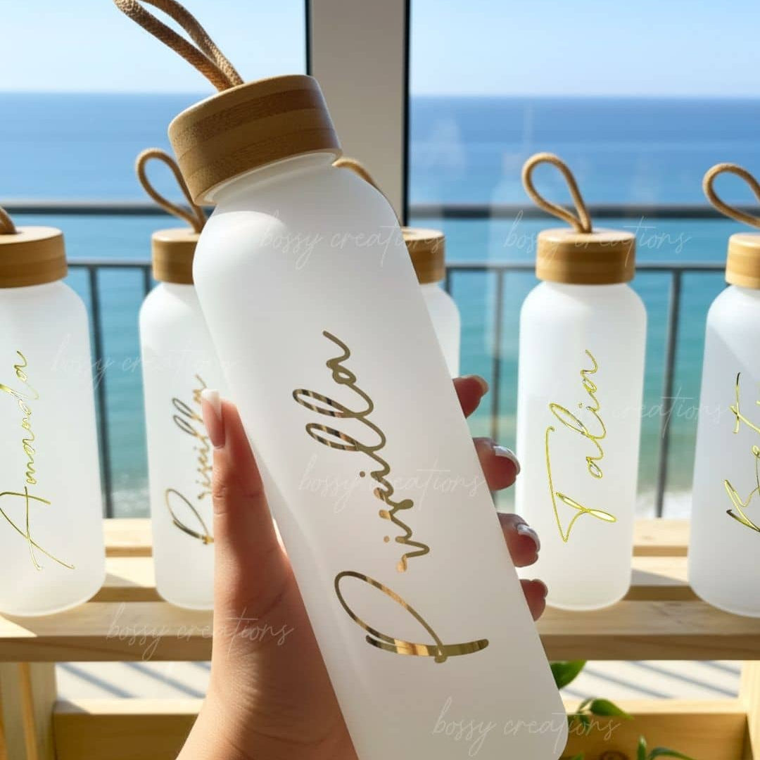 glass water bottles with personalized gold text and wooden caps held by a hand, with a scenic background for bridesmiad gifts.
