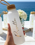 White glass water bottle with personalized gold text held by a hand, set against a scenic ocean backdrop during a beach bachelorette.
